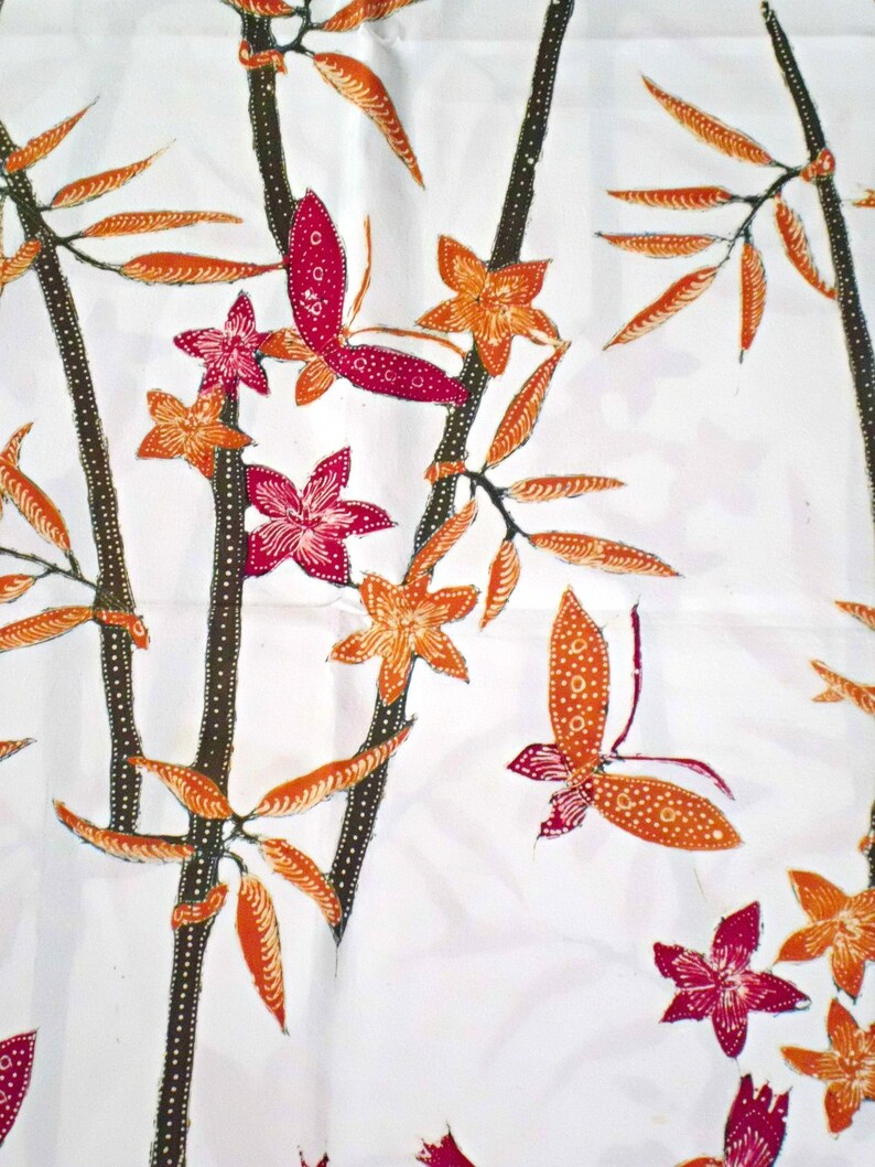 May include: White fabric with a repeating pattern of orange and brown leaves, pink and red flowers, and orange and red butterflies. The pattern is reminiscent of a traditional Indonesian batik design.