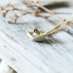 Könnte beinhalten: Eine zarte Halskette mit einem kleinen, cremefarbenen Papierboot-Anhänger. Der Anhänger hängt an einer feinen, goldfarbenen Kette. Die Halskette liegt auf einer verwitterten, hellen Holzoberfläche, mit getrockneten floralen Akzenten im Hintergrund.