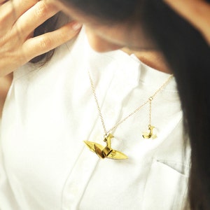 K&ouml;nnte beinhalten: Eine goldene Halskette mit zwei Origami-Kranich-Anh&auml;ngern. Der gr&ouml;&szlig;ere Kranich befindet sich an einer l&auml;ngeren Kette und der kleinere Kranich an einer k&uuml;rzeren Kette.