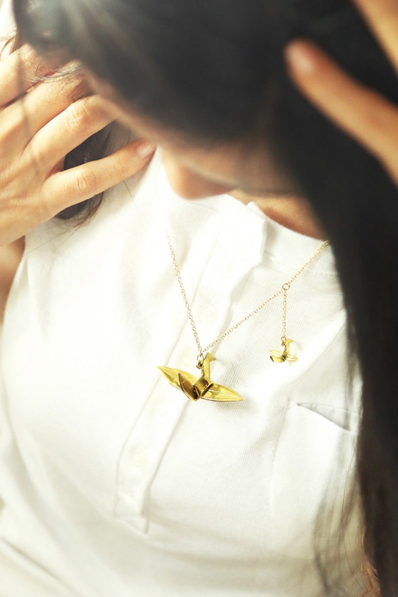 K&ouml;nnte beinhalten: Eine goldene Halskette mit zwei Origami-Kranich-Anh&auml;ngern. Der gr&ouml;&szlig;ere Kranich befindet sich an einer l&auml;ngeren Kette und der kleinere Kranich an einer k&uuml;rzeren Kette.
