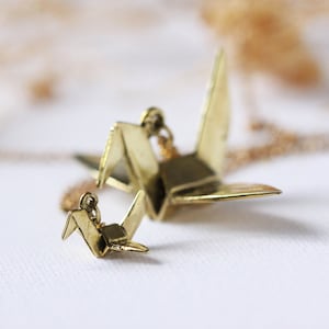 K&ouml;nnte beinhalten: Eine goldene Halskette mit zwei Origami-Kranich-Anh&auml;ngern. Der gr&ouml;&szlig;ere Kranich befindet sich an einer Kette und der kleinere Kranich ist an dem gr&ouml;&szlig;eren Kranich befestigt.
