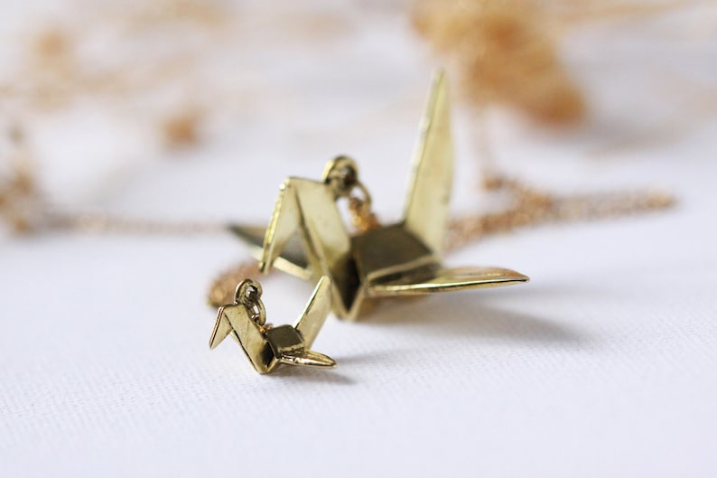 K&ouml;nnte beinhalten: Eine goldene Halskette mit zwei Origami-Kranich-Anh&auml;ngern. Der gr&ouml;&szlig;ere Kranich befindet sich an einer Kette und der kleinere Kranich ist an dem gr&ouml;&szlig;eren Kranich befestigt.