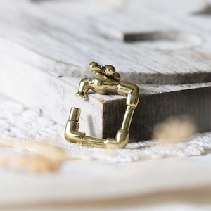 May include: A gold-toned metal faucet charm in the shape of a square. The faucet has a small handle and a spout. The charm is on a white wooden surface.