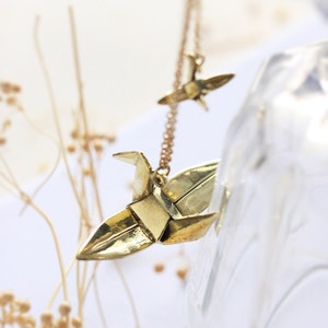 K&ouml;nnte beinhalten: Eine goldene Halskette mit zwei Origami-Kranich-Anh&auml;ngern. Die Kraniche sind aus Metall und haben eine gl&auml;nzende Oberfl&auml;che.