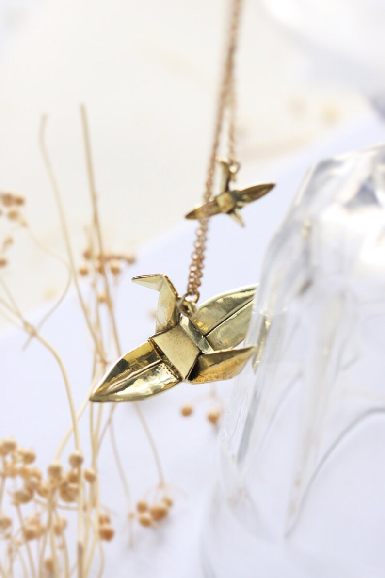K&ouml;nnte beinhalten: Eine goldene Halskette mit zwei Origami-Kranich-Anh&auml;ngern. Die Kraniche sind aus Metall und haben eine gl&auml;nzende Oberfl&auml;che.