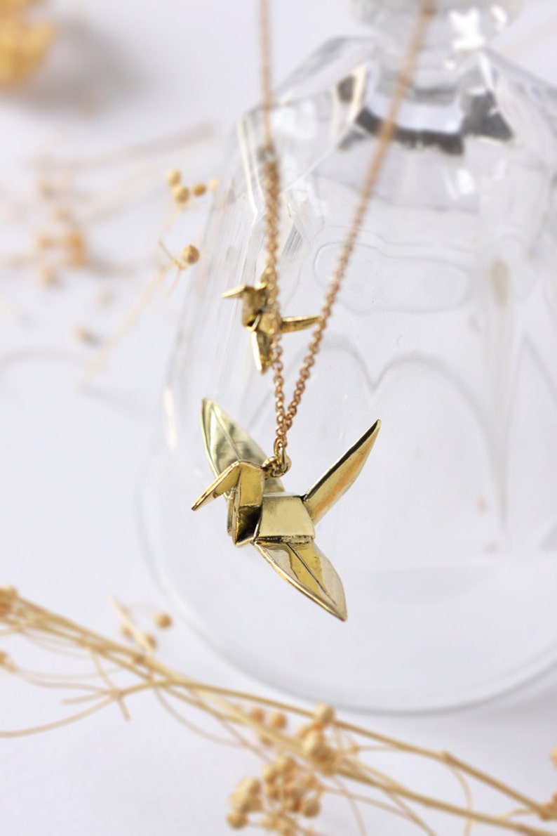 K&ouml;nnte beinhalten: Eine goldene Halskette mit zwei Origami-Kranich-Anh&auml;ngern. Die Kraniche sind im Flug und die Kette h&auml;ngt an einem Glasbeh&auml;lter.