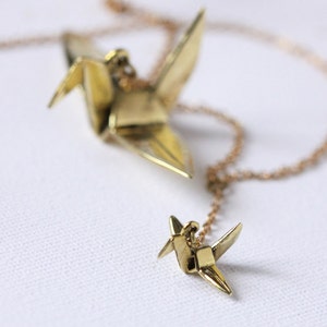 K&ouml;nnte beinhalten: Eine goldene Halskette mit zwei Origami-Kranich-Anh&auml;ngern. Der gr&ouml;&szlig;ere Kranich befindet sich oben an der Kette und der kleinere Kranich unten.