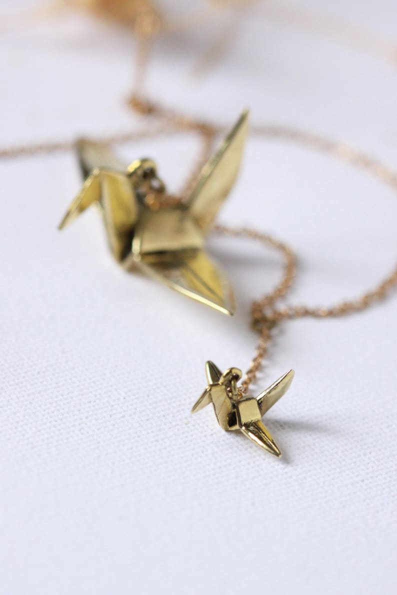 K&ouml;nnte beinhalten: Eine goldene Halskette mit zwei Origami-Kranich-Anh&auml;ngern. Der gr&ouml;&szlig;ere Kranich befindet sich oben an der Kette und der kleinere Kranich unten.