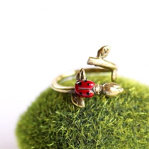 Könnte beinhalten: Ein goldfarbener Ring mit einem Marienkäfer-Design. Der Ring hat einen zarten Zweig mit Blättern und einen roten Marienkäfer mit schwarzen Punkten.