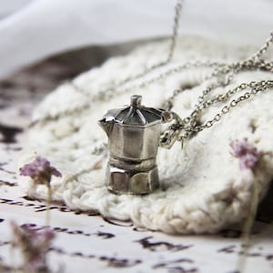 May include: A silver-coloured pendant necklace with a miniature coffee pot charm. The charm is a detailed replica of a stovetop espresso maker. The necklace rests on a white crocheted doily, with dried flowers in the background.