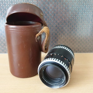 May include: A vintage camera lens with a black and silver design, next to a brown leather case with a handle. The lens has a series of markings and numbers around the barrel. The case is cylindrical with a hinged lid.