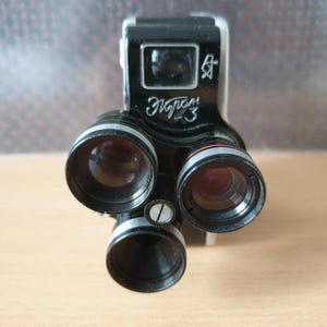 Puede incluir: Una cámara vintage en blanco y negro con tres lentes y el texto cirílico "Экрам-3". La cámara tiene un cuerpo rectangular con un visor y un anillo de lente plateado. Las lentes son oscuras y redondas.