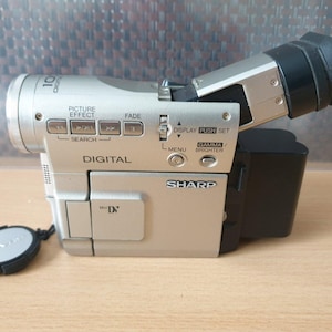 May include: Silver Sharp digital camcorder with a black eyepiece and lens cap. The camcorder has buttons for picture effects, fade, search, and menu. The word "DIGITAL" is printed on the front.