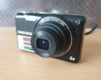 Könnte beinhalten: Schwarze Samsung Digitalkamera mit Zoomobjektiv. Die Kamera hat eine rechteckige Form mit einem Objektiv, das aus der Vorderseite herausragt. Das Objektiv ist von einem silbernen Ring umgeben. 5x Zoom.