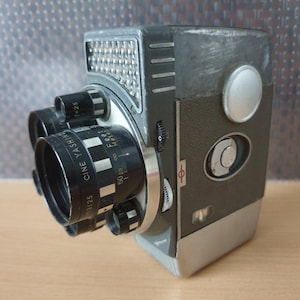 以下が含まれることがあります： 複数のレンズと内蔵ライトを備えた、ヴィンテージのヤシカシネカメラ。 カメラ本体はグレーと黒の混合で、シルバーのアクセントが付いています。 レンズには黒とシルバーのディテールがあり、フィートとメートルの測定値が表示されています。