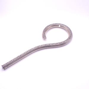 May include: A silver-coloured, curved metal object with a textured surface. The object has a hook-like shape, with a rounded top and a long, slightly curved stem. The texture appears to be a pattern of small, closely spaced dots.
