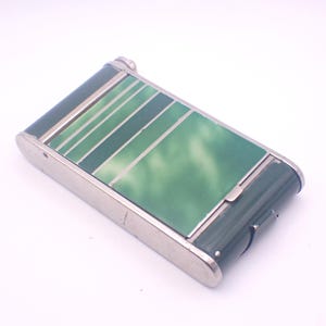 May include: Rectangular, vintage cigarette case with a green and silver design. The case has a dark green body with a silver frame and a green and white marbled top. The top features diagonal green stripes.