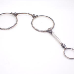 Op de afbeelding: Antieke zilverkleurige bril met ronde glazen en een handvat. De monturen hebben ingewikkelde decoratieve patronen. Het handvat heeft een kleine ring aan het uiteinde. De bril staat tegen een witte achtergrond.