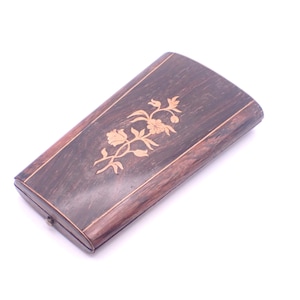 Puede incluir: Una caja rectangular de madera con un acabado marrón oscuro y una incrustación floral decorativa en un tono marrón más claro. La caja tiene una tapa con bisagras y un pequeño cierre. El diseño general es elegante y vintage.