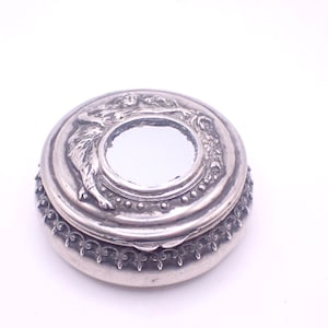 May include: A small, round, silver-toned trinket box with a mirrored center. The lid features a detailed relief of a figure and floral elements. The box has a decorative beaded and patterned edge.