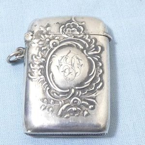Può includere: Un portafiammiferi d'argento con un ornato motivo floreale e a volute. La custodia rettangolare presenta un coperchio incernierato e un cartiglio ovale decorativo con iniziali incise. Un piccolo anello è attaccato al lato.