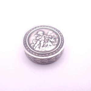 May include: A small, round, silver-toned box with a detailed relief design. The lid features a central image of a cherub with a bow and arrow, surrounded by a decorative border. The box's side has a repeating leaf pattern.