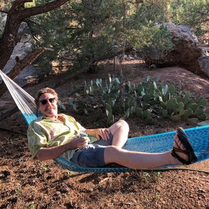 May include: A man wearing sunglasses and a green shirt relaxes in a blue and white hammock. He is holding a can of beverage and is surrounded by desert vegetation.