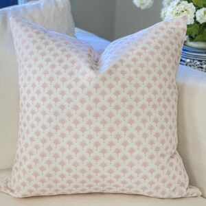 May include: A square decorative pillow with a white background and a repeating pink floral pattern. The pillow is placed on a white sofa, with a white pillow behind it. A vase of white flowers is visible in the background.