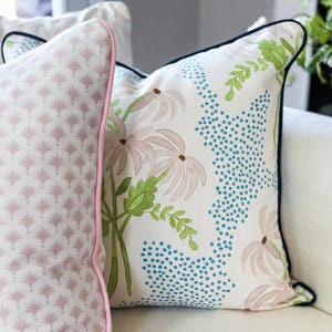 May include: Two square throw pillows. The left pillow features a repeating light pink fan-like pattern with pink piping. The right pillow has a white background with a floral design of light pink flowers, green leaves, and blue dotted accents with dark piping.