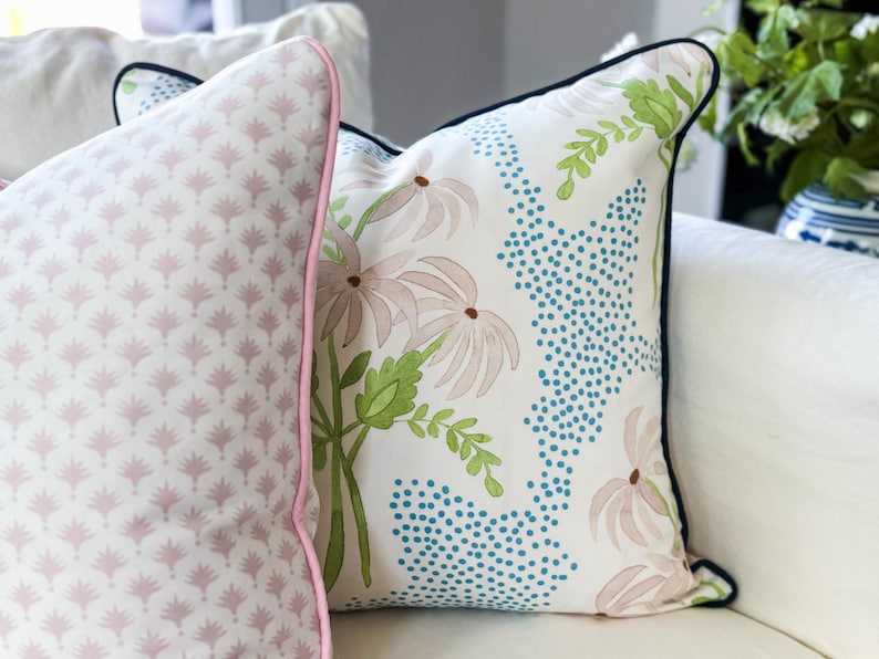 May include: Two square throw pillows. The left pillow features a repeating light pink fan-like pattern with pink piping. The right pillow has a white background with a floral design of light pink flowers, green leaves, and blue dotted accents with dark piping.