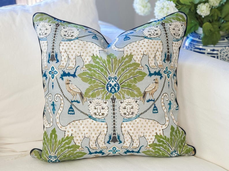 May include: Square decorative pillow with a light blue background featuring a repeating pattern of white spotted leopards, birds, and green palm trees. The pillow has a black border and is placed on a white surface.
