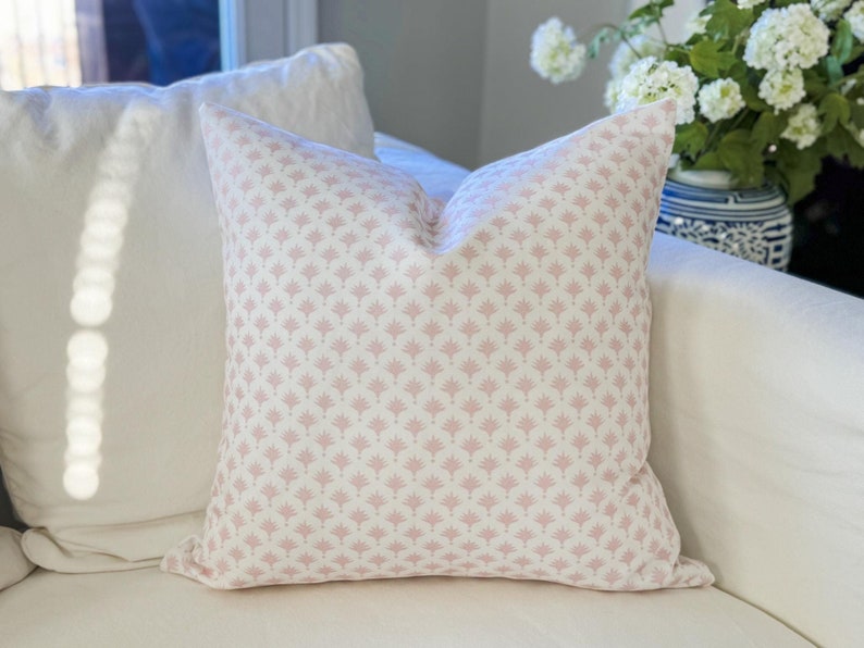 May include: A square decorative pillow with a white background and a repeating pink floral pattern. The pillow is placed on a white sofa, with a white pillow behind it. A vase of white flowers is visible in the background.