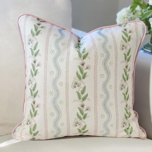May include: A square throw pillow featuring a cream-colored fabric with a vertical pattern of green leaves, pink flowers, and wavy blue lines. The pillow is finished with a solid pink piped edge that outlines the entire square shape of the cushion.