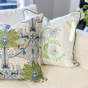 May include: Two decorative throw pillows with unique patterns. One features a light blue background with white leopards, green palm trees, and birds. The other pillow has a white background with a green floral design and a green border.