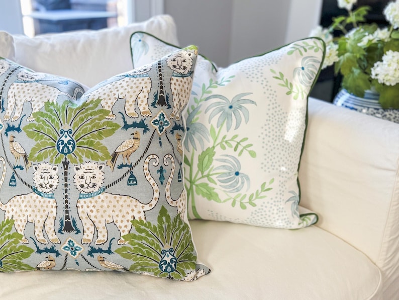 May include: Two decorative throw pillows with unique patterns. One features a light blue background with white leopards, green palm trees, and birds. The other pillow has a white background with a green floral design and a green border.
