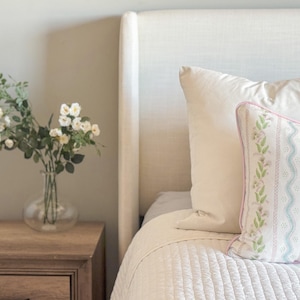May include: A bedroom scene featuring a bed with white bedding and pillows. A decorative pillow with a floral pattern and pink trim rests against a white pillow. A wooden nightstand holds a glass vase with white flowers.