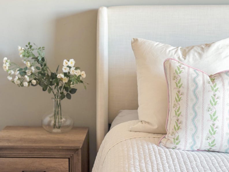 May include: A bedroom scene featuring a bed with white bedding and pillows. A decorative pillow with a floral pattern and pink trim rests against a white pillow. A wooden nightstand holds a glass vase with white flowers.
