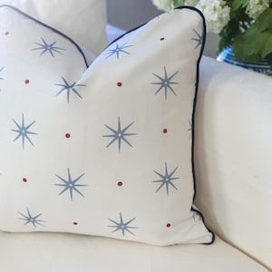 May include: A square throw pillow with a white base featuring a repeating pattern of light blue eight-pointed starbursts and small red dots. The pillow is finished with a dark navy blue piped edge along the perimeter of the fabric cover.