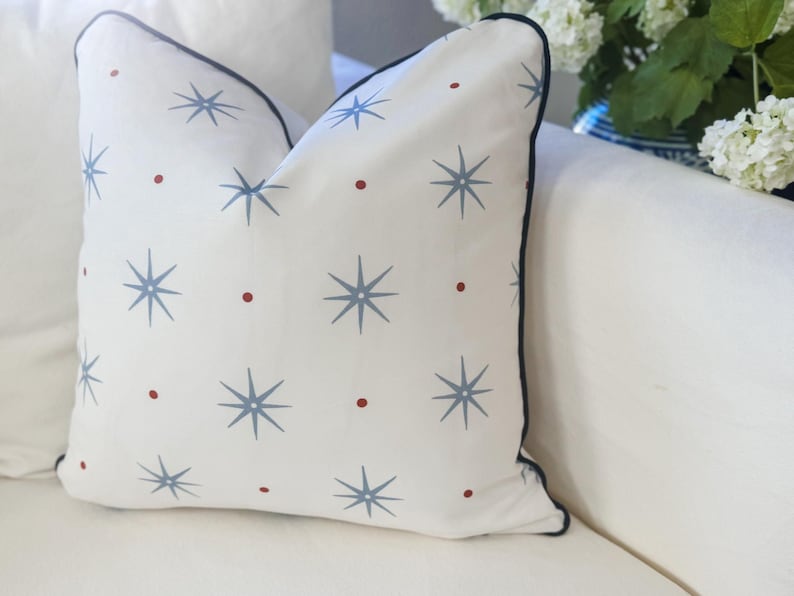 May include: A square throw pillow with a white base featuring a repeating pattern of light blue eight-pointed starbursts and small red dots. The pillow is finished with a dark navy blue piped edge along the perimeter of the fabric cover.