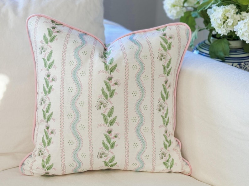 May include: Square decorative pillow with a white background and a repeating floral pattern in green, pink, and light blue. The pillow has a pink border and is placed on a white surface.