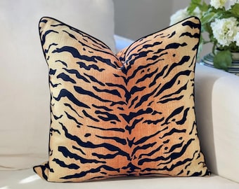 May include: A square throw pillow featuring a tiger stripe pattern in shades of orange and black. The fabric has a visible woven texture, and the edges of the pillow are finished with a thin black piping trim along the entire perimeter.