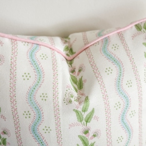 May include: A decorative pillow with a floral pattern. The pillow features a white background with vertical stripes of green leaves and pink flowers. A light blue wavy stripe runs down the center. The pillow is trimmed with a pink piping.