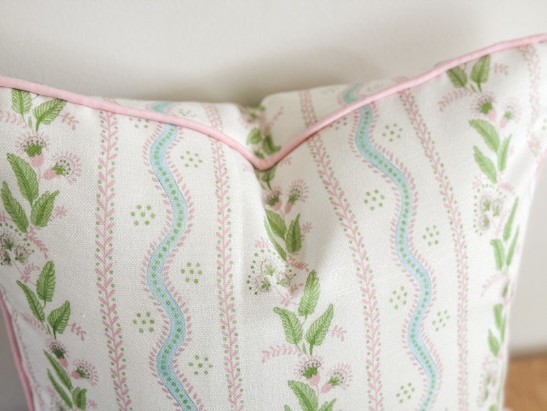 May include: A decorative pillow with a floral pattern. The pillow features a white background with vertical stripes of green leaves and pink flowers. A light blue wavy stripe runs down the center. The pillow is trimmed with a pink piping.