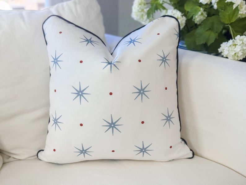 May include: A square throw pillow with a white base featuring a repeating pattern of light blue eight-pointed stars and small red dots. The pillow is finished with a dark blue piped edge that outlines the entire square shape of the cushion.