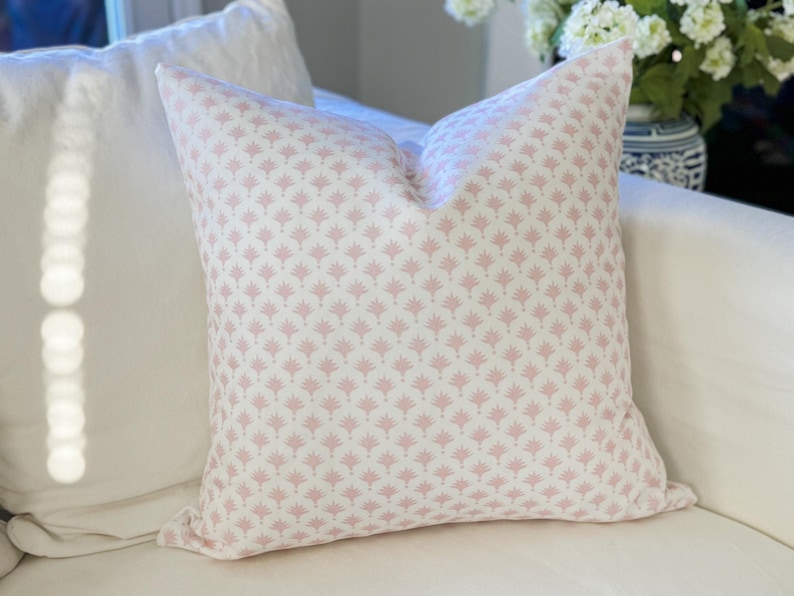 May include: A square throw pillow featuring a repeating pattern of small, light pink fan-shaped motifs printed on a solid white fabric background. The pillow has a clean, structured shape with visible seams along the edges and a soft, plush appearance.