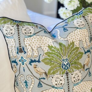 May include: Decorative throw pillow with a light blue background featuring a repeating pattern of white tigers with brown spots, green palm trees, and birds. The pillow has a black border and is placed on a white surface.