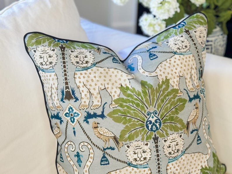 May include: Decorative throw pillow with a light blue background featuring a repeating pattern of white tigers with brown spots, green palm trees, and birds. The pillow has a black border and is placed on a white surface.