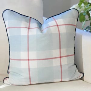 May include: A square throw pillow featuring a light blue and white plaid pattern with thin red intersecting lines. The pillow is finished with a dark navy blue piping along all four edges, providing a defined border to the soft fabric design.
