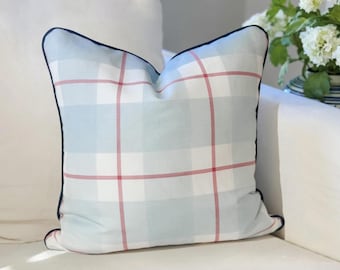 May include: A square throw pillow featuring a light blue and white plaid pattern with thin red intersecting lines. The pillow is finished with a dark navy blue piping along all four edges, providing a defined border to the soft fabric design.