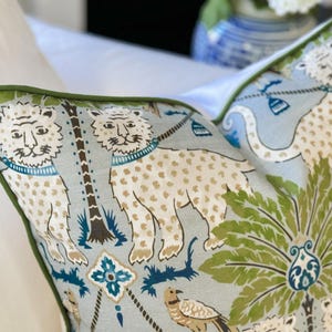 May include: Decorative pillow with a light blue background and a repeating pattern of lions, palm trees, and other botanical elements. The lions are white with tan spots and blue collars. The pillow has a green border.